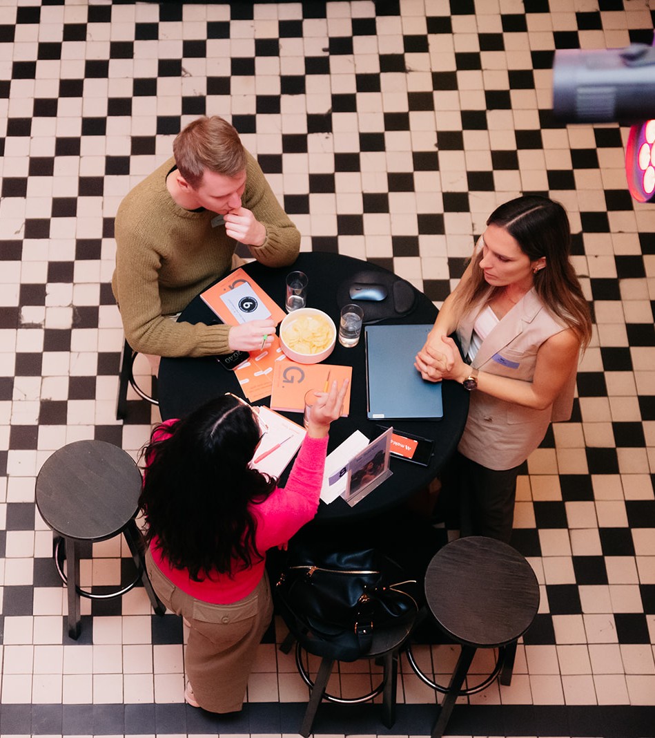 Three people in conversation around a small table at a networking event, seen from above, with a laptop and papers between them